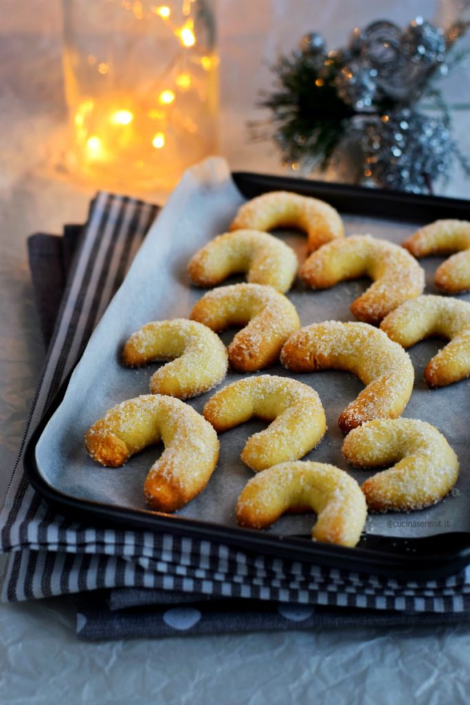 vanillikipferl biscotti alla vaniglia a forma di mezzaluna  messi su una teglia ricoperta di carta forno adagiata su canovacci a righe bianco e grigio con dietro lucine e decorazioni di natale