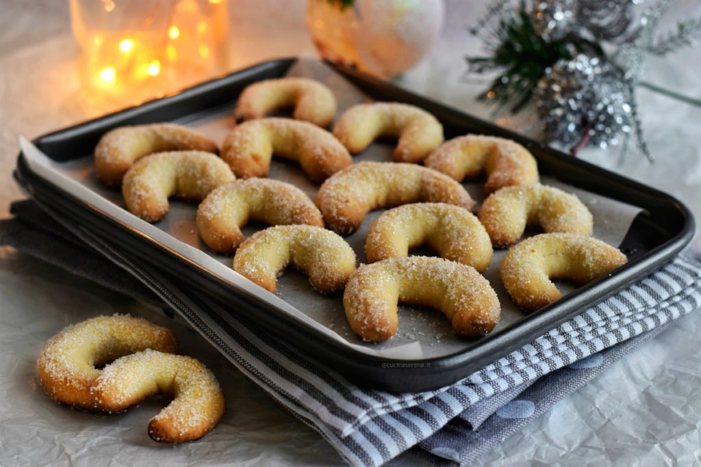 biscotti vanillekipferl a forma di mezzaluna ricoperti di zucchero adagiati su una teglia da forno posta su strofinacci color grigio sullo sfondo lucine di natale  e palline di natale