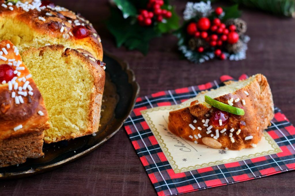 Fetta di ciambella Rescon de Reyes dolce lievitato spagnolo decorato con ciliege, cedro e arance candite, mandorle e granella di zucchero messa su un tovagliolino natalizio. A sinistra su un vassoio si intravede metà della ciambella tagliata con un altra fetta accostata al dolce intero