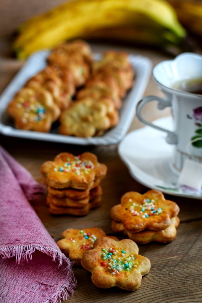 biscotti a forma di fiore decorati con diavolini, (ovvero confettini colorati) impilati uno sugli altri. Tre file di 3 e due biscotti impilati e altri due di poco sovrapposti sul tavolo. sullo sfondo un vassoio pieno di biscotto, due tazze da te e sulla sinistra un tovagliolo rosa di lino sfrangiato