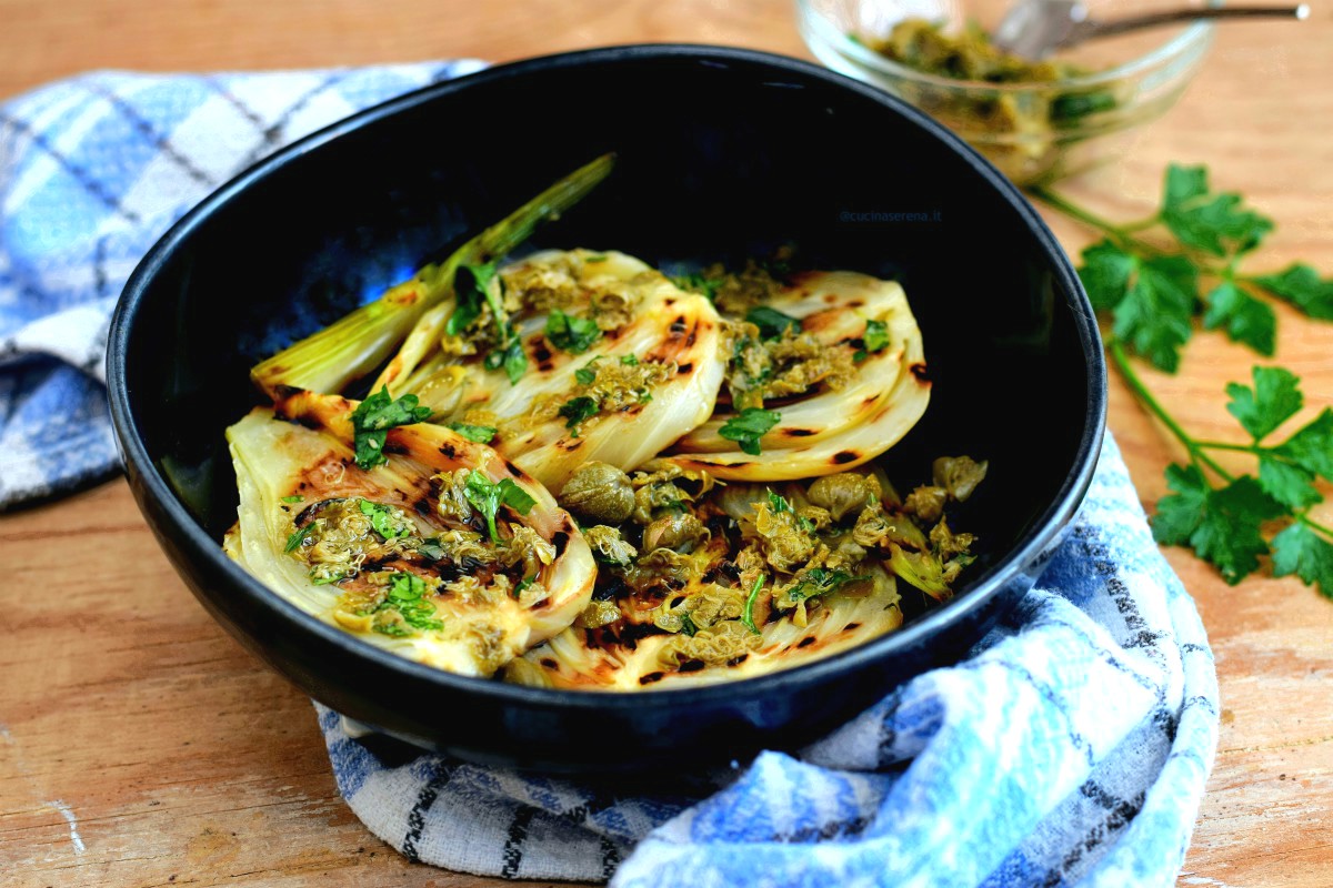 finocchi grigliati su piastra conditi con una salsa fatta di capperi tritati limone e olio. I finocchi sono posti in un piatto concavi blu dalla sfumature celesti. Sotto il piatto c'è un canovaccio bianco a righe blu  celesti e blu. Dietro al piatto in alto sulla destra si intravede una ciotola con il battuto di capperi e un cucchiaino per servirla e due rametti di prezzemolo, ingrediente presente nella salsa di capperi.