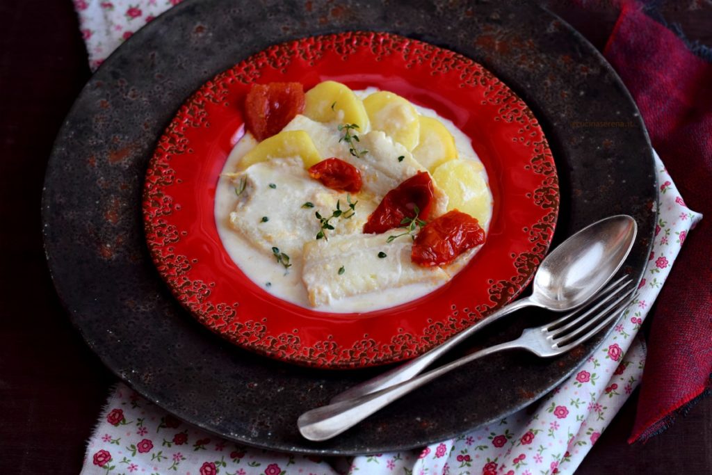 Piatto rosso con sottopiatto vintage color peltro con venature rossastre. Nel piatto di ceramica c'è il merluzzo in una salsa di latte con fettine di patate pezzi di pomodoro e foglioline di timo