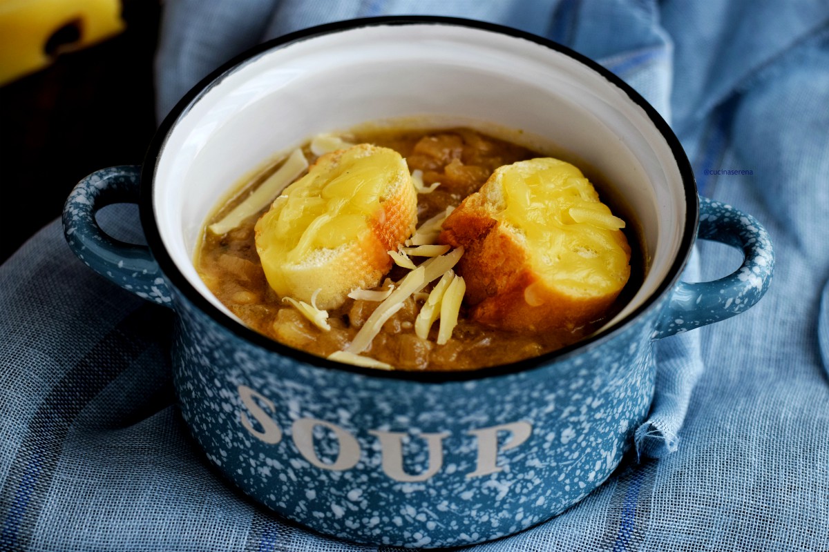 Soupe à l'oignon - zuppa di cipolle  francese in un pentolino celeste maculato fuori con una scritta bianca SOUP e smaltato all'interno bianco con bordi neri. Nel pentolino c'è la zuppa di cipolle due fette di baguette abbrustolite con groviera fusa sopra e scaglie di groviera per decorare. La zuppa viene infornata con i crostini prima di essere servita in modo che il formaggio si fonda