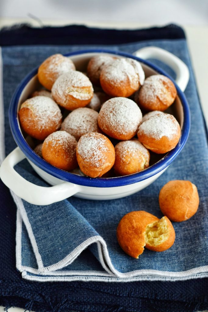 Castagnole con mascarpone fritte, nell'immagine sono raccolte in una ciotolina bianca con bordi blue e due manicia all'esterno. La ciotola è adagiata su una stoffa jeans e di lato sul tovagliolo ci sono due castagnole di cui una aperta a metà.