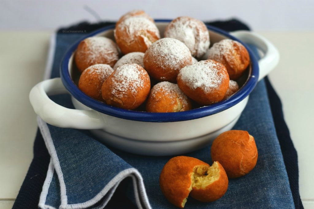 Castagnole al mascarpone fritte ricoperta di zuccchero a velo in unca ciotolaa bianca adagiata su un tovagliolo di jeans. Accanto due castagnole di cui una aperta dalla quale si intravede l'interno