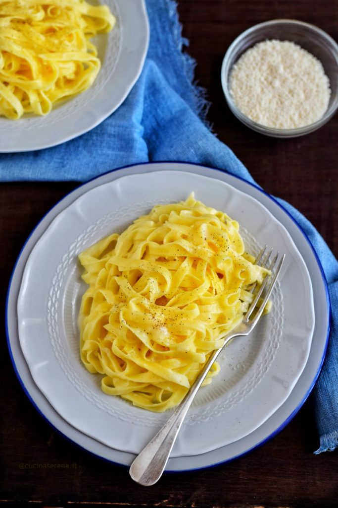 Su un tavolo di legno scuro c'è un piatto bianco su un sottopiatto con bordo blu adagiato su un tovagliolo di lino celeste sfrangiato. Nel piatto ci sono le fettucine Alfredo, pasta in bianco mantecata con abbondante burro e parmigiano con sopra una spolverata di pepe. Il piatto è posizionato nel centro in basso a sinistra, più in alto sulla destra una ciotola di parmigiano grattugiato a sinistra invece si intravete un quarto di un altro piatto con le fettuccine.