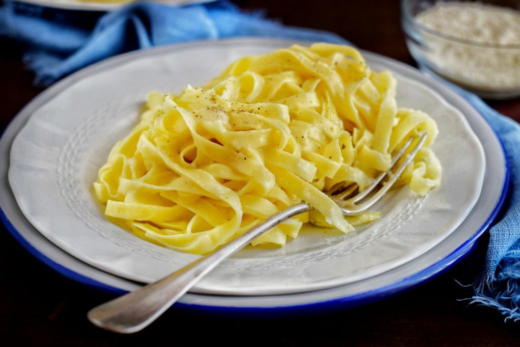 Fettuccine Alfredo spolverizzate di pepe in un piatto bianco con una posata di lato a destra che infilsa la pasta. il piatto è adagiato su un sottopiatto con bordo blu, dietro una stoffa di lino sfrangiata 