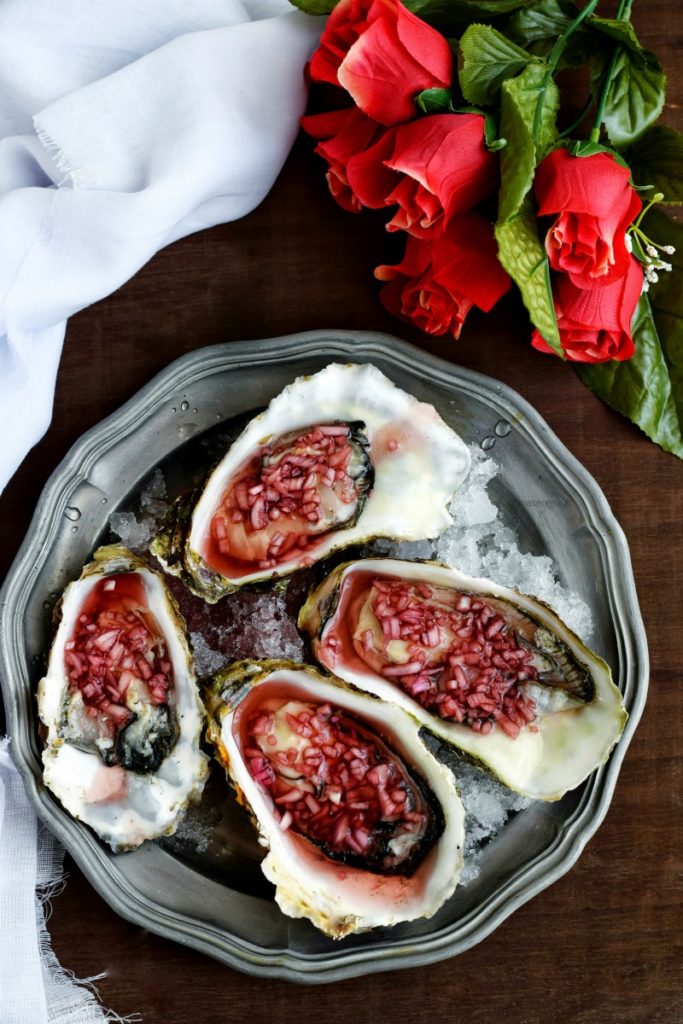 Ostriche condite con dressing mignonette fatto con scalogno, vino, aceto e pepe. Le ostriche sono adagiate su un vassoio in peltro con ghiaccio tritato, sullo sfondo un mazzo di rose e un drappo bianco di tela. Immagine zenitale come fosse vista per intero dall'alto al basso 