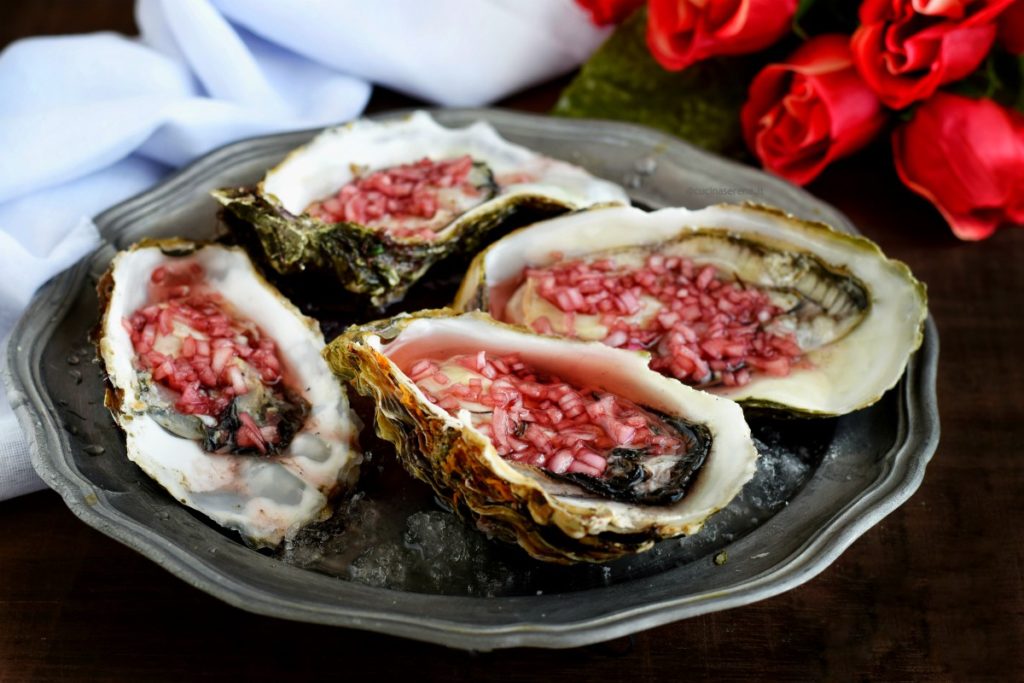 Ostriche condite con dressing mignonette fatto con scalogno, vino, aceto e pepe. Le ostriche sono adagiate su un vassoio in peltro con ghiaccio tritato, sullo sfondo un mazzo di rose e un drappo bianco di tela. Immagine orizzontale visione frontale