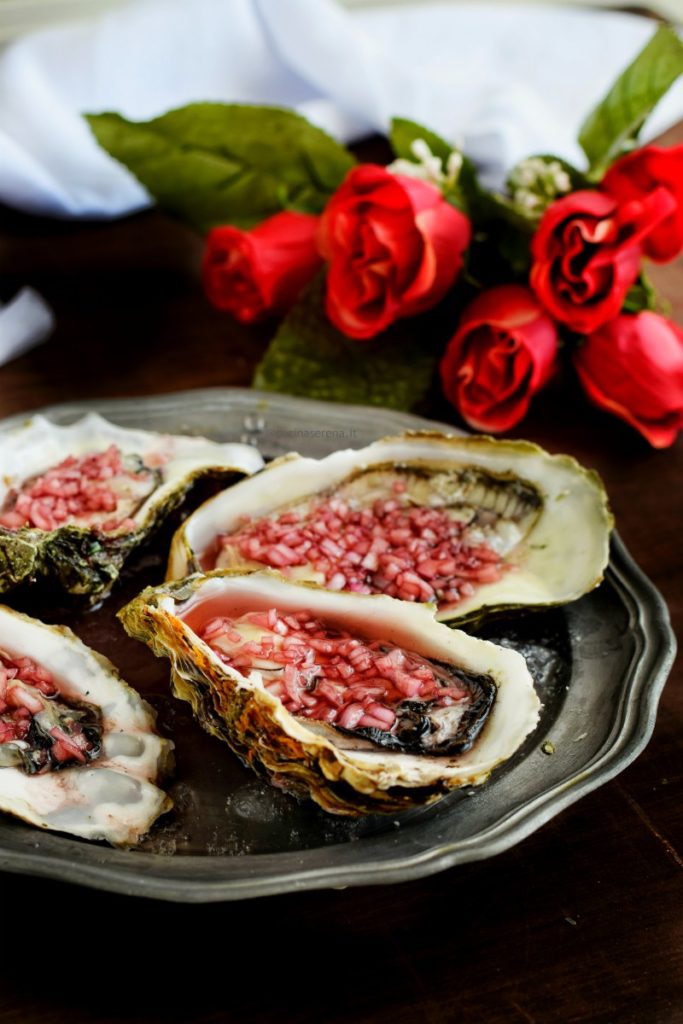 Ostriche condite con dressing mignonette fatto con scalogno, vino, aceto e pepe. Le ostriche sono adagiate su un vassoio in peltro con ghiaccio tritato, sullo sfondo un mazzo di rose e un drappo bianco di tela