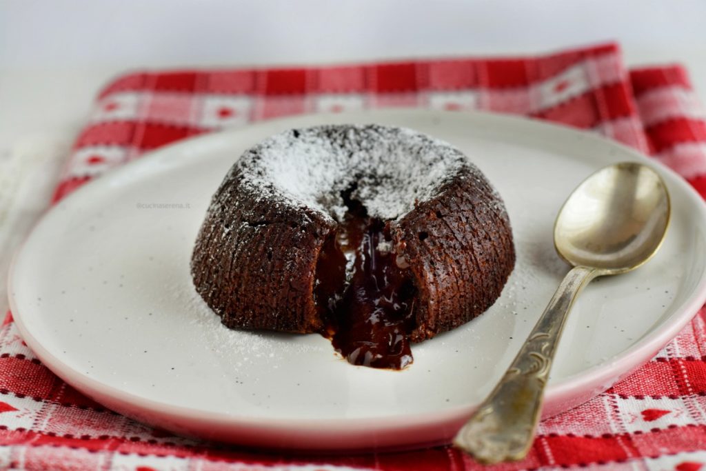 tortino al cioccolato fondente dalla forma rotonda messo in un piattino con un cucchiaino di lato. Il tortino è aperto e dalla fessura fuoriesce del cioccolato liquido