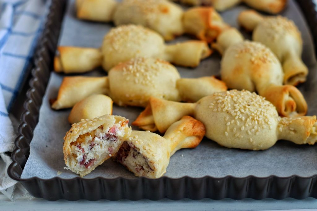 Caramelle di pasta matta tagliata in due con ripieno di ricotta e salame visibile. Sullo sfondo altre caramelle di pasa matta intere su una teglia da forno