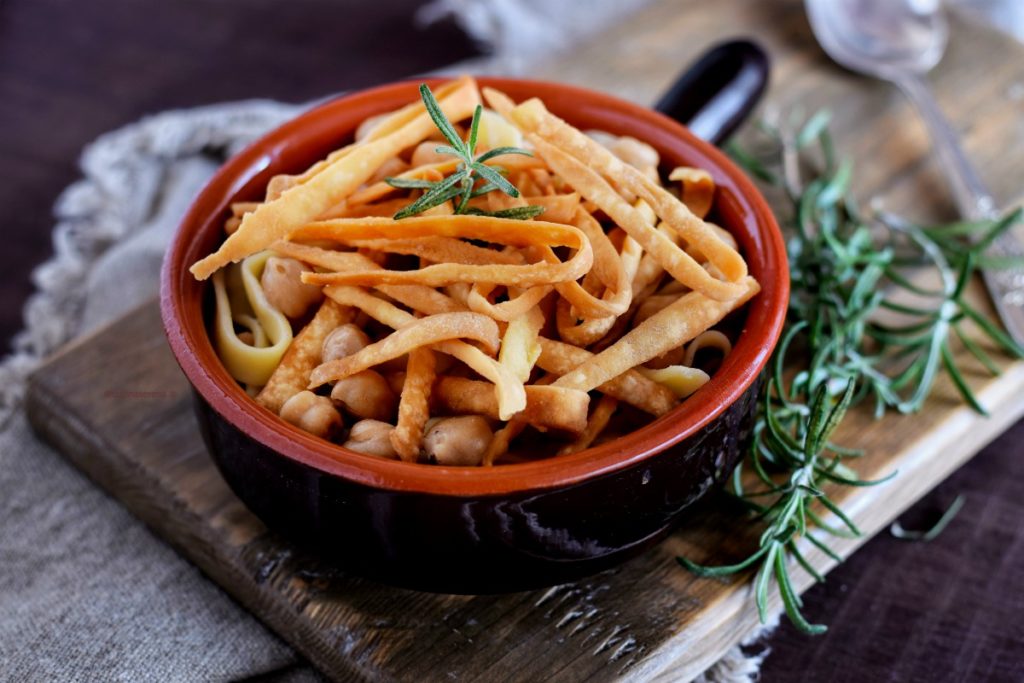 Ciceri e tria ricetta salentina servita un ciotola di coccio con manico, adagiata su un tagliere rustico sotto un telo grezzo. Il paiatto presenta la tipica pasta fritta oltre a quella cotta nella zuppa di ceci