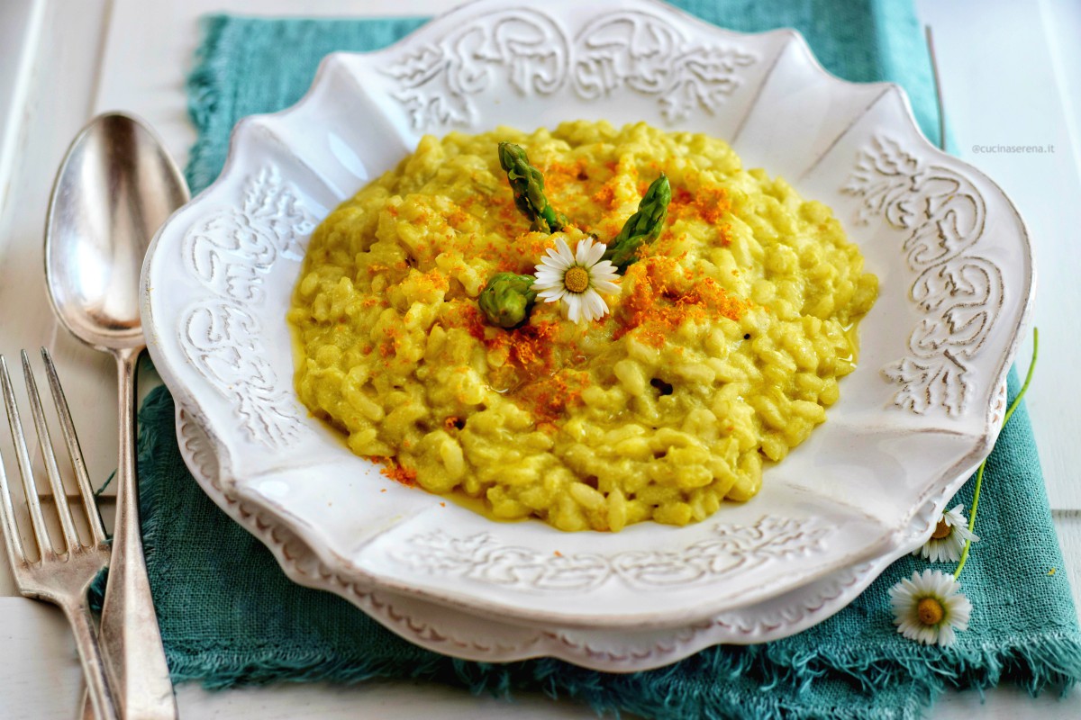 Risotto asparago e arancia servito su un piatto bianco serigrafato, sotto al quale c'è un sottopiatto adagiato su un tovagliolo verde di lino, a sinistra cucchiaio e forchetta