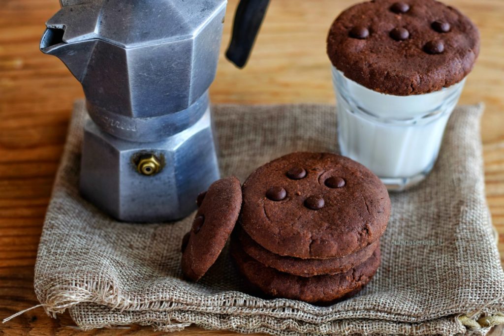 Biscotti al cioccolato al microonde in pochi minuti. Nella foto sono adagiati su una stoffa di iuta di lato la macchinetta del caffè e sull'altro lato un bicchiere con il latte sul quale è deposto un biscotto come fosse un tappo