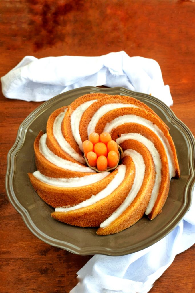 bundt cake carote, zucchina e polvere di liquirizia presentata su un alzatina al centro della ciambella con foro un mazzetto di carotine baby legate da uno spago