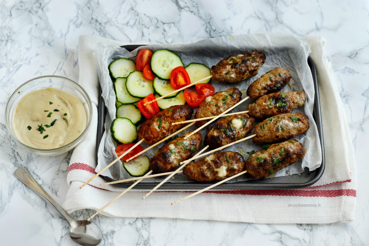 Kofta secondo piatto di carne di origine medio orientale nella foto servito su una teglia ricoperta di carta forno su cui sono adagiate le kofte in versione spiedino con accanto pomodori e cetrioli a fette