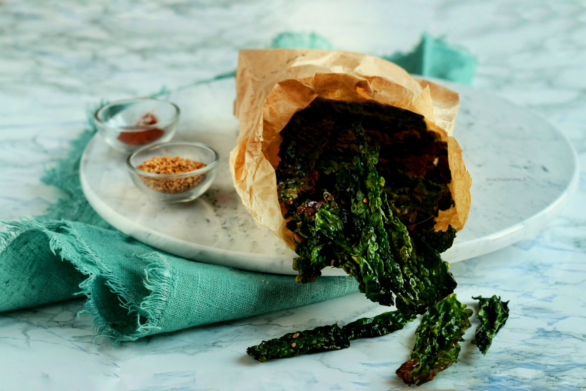 vegan chips di cavolo nero al forno. ricetta facile e veloce nella foto prensentate in un sacchetto di carna come fossero patatine con accanto due ciotoline piccole con sesamo e paprika a indicarne le varianti alle versione semplice con sola olio e sale