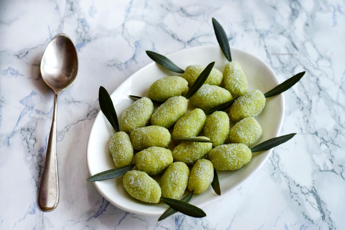 olivette in pasta di mandorel nella foto servite in un piatto bianco con foglie di ulivo come decorazione e un cucchiaio per prenderle accanto al piatto