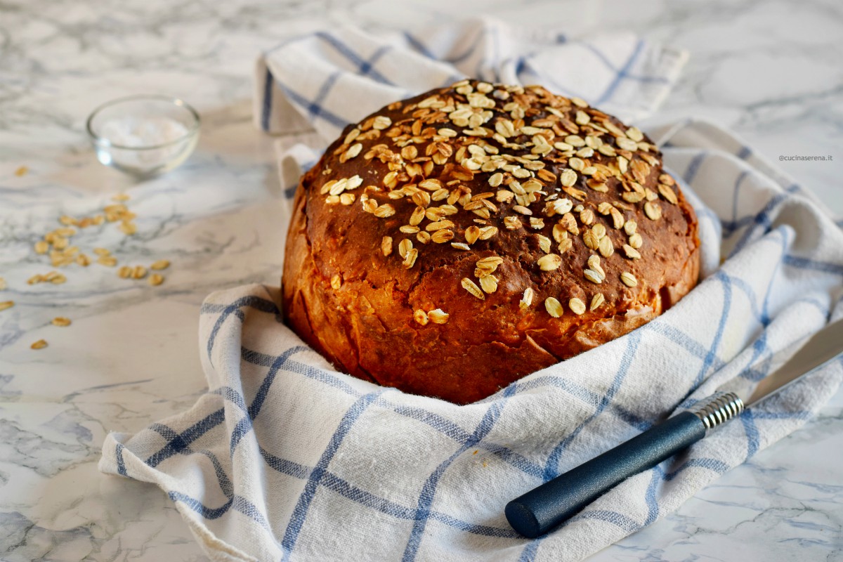 soda bread nella foto avvolto in un canovaccio bianco con righe a quadri blu, il pane è ricoperto di fiocchi di avena sullo sfondo si vede il bicarbonato e i fiocchi di avena