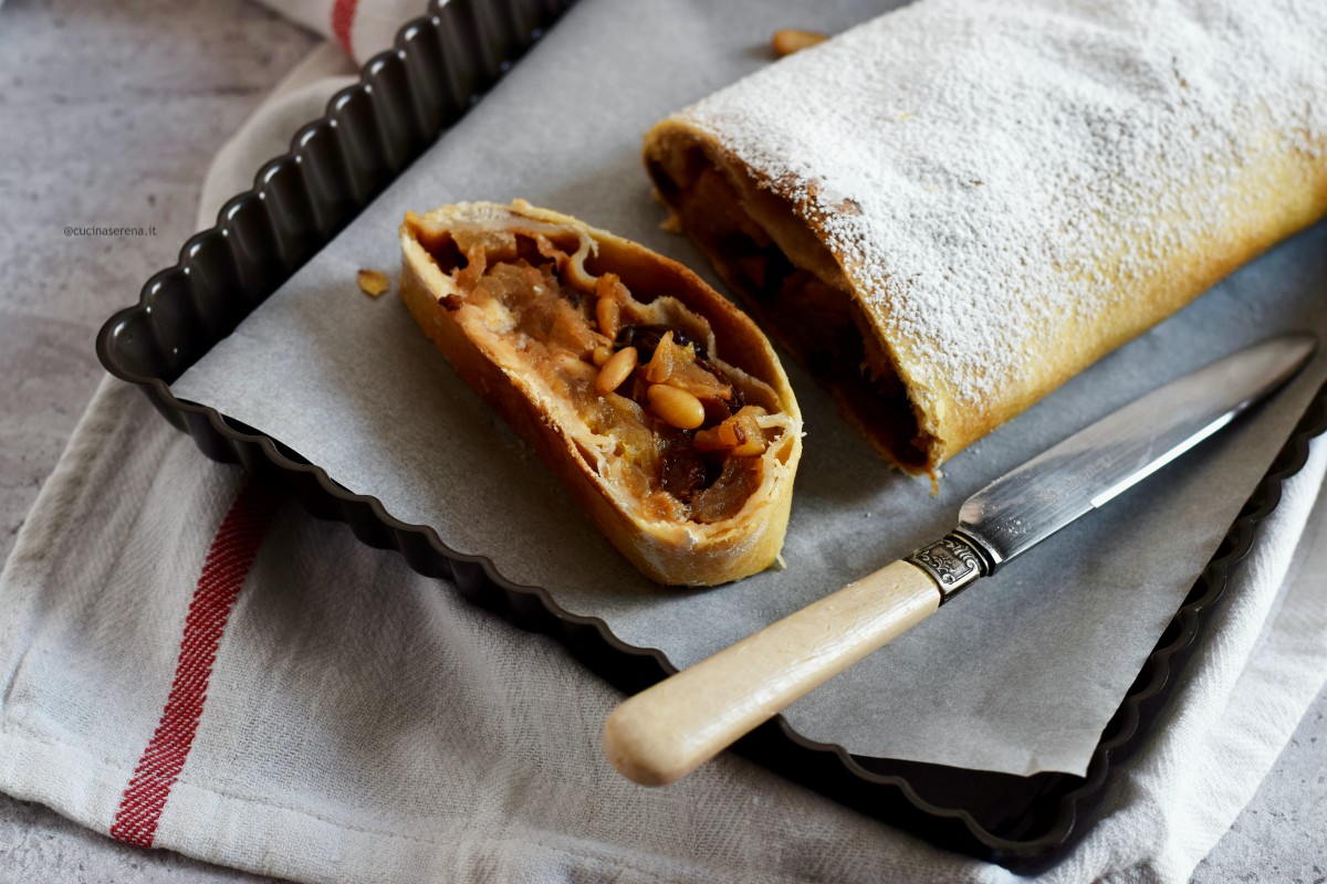 Strudel di mele nella foto una fetta tagliata adagiata su una teglia con carta forno. Dalla fetta di vedono tutti gli ingredienti che compongono lo strudel