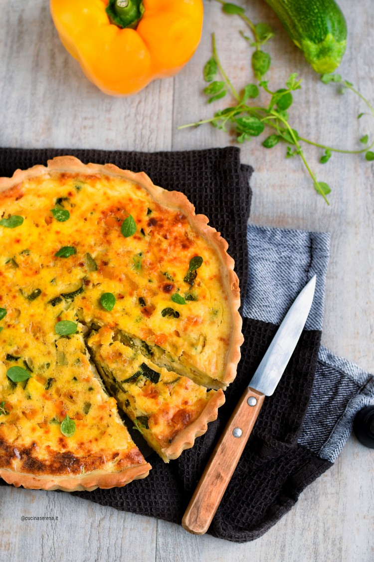 torta rustica di pasta matta con zucchine e peperoni - nella foto la torta adagiata su un canovaqccio con una fetta tagliata sullo sfondo le verdure della torta e la maggiorana