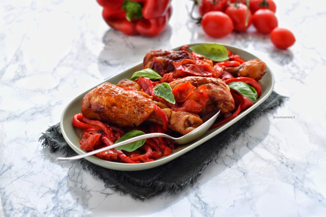 Pollo con i peperoni alla romana: pollo a pezzi cotto in pentola con pomodori al quale vanno poi aggiunti i peperoni, nella foto servito in un vassoio. Sullo sfondo peperoni e pmodori