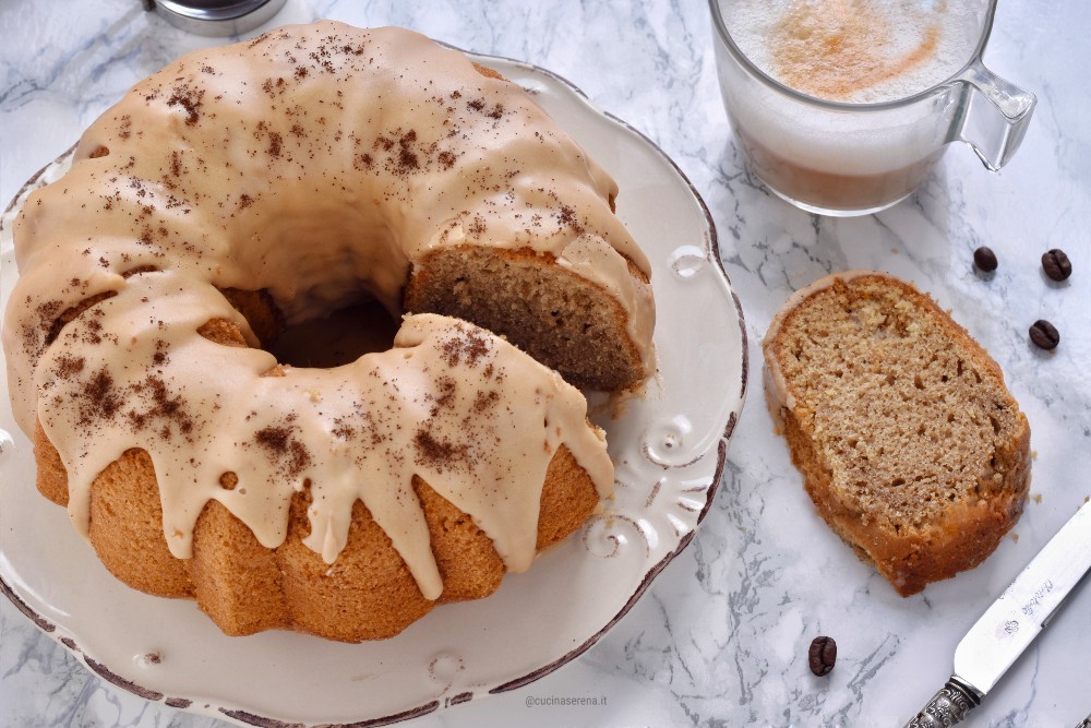 Torta al cappuccino con glassa al caffè presentata in un piatto, tagliata con una fetta accanto al piatto con il cappuccino sullo sfondo